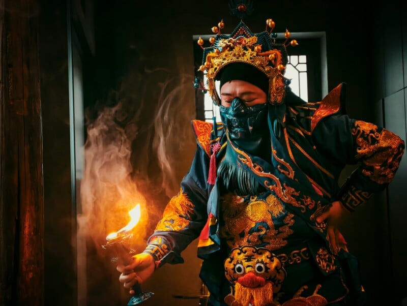 A person in ornate traditional Chinese opera costume and headpiece holds a flaming object indoors, with dramatic lighting and smoke swirling around them.