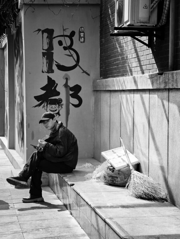 A person in a cap sits on a low wall, eating food, beside a broom and a box. Behind them, a wall features large Chinese characters and an illustrated face. The image is in black and white.