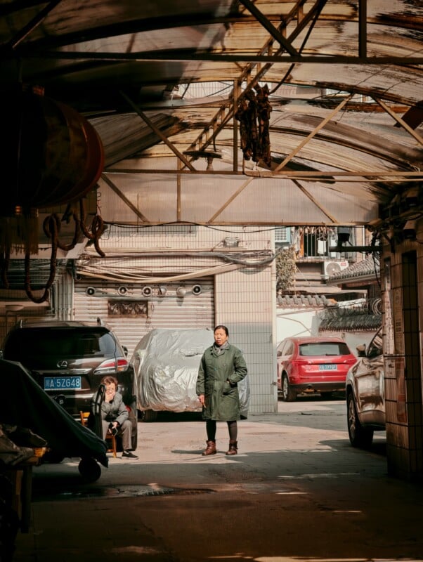 A man in a green coat stands in a covered alleyway, while an older person sits nearby. Parked cars and residential buildings are visible in the background on a sunny day.