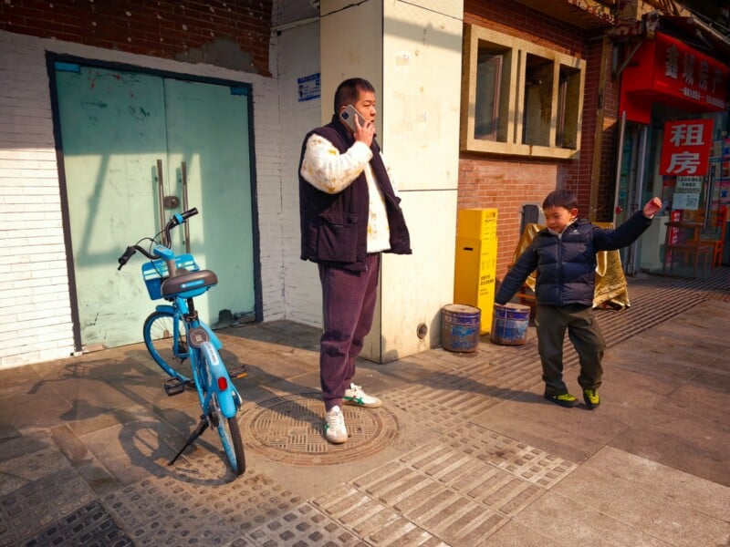 A man talks on his phone next to a blue bicycle, while a young boy in a puffy jacket stands nearby with his arm outstretched on a sunny sidewalk in front of a brick building.