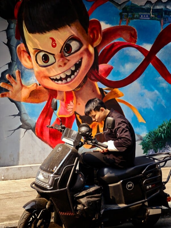 A man sits on a black scooter in front of a colorful mural of a mischievous cartoon character with large eyes, a red cape, and a playful expression. The mural appears as if the character is bursting through the wall.