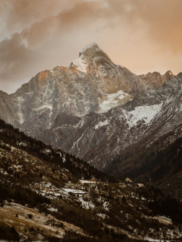 Snow-capped mountain peak is illuminated by soft orange sunset light, with rugged slopes and a forested valley below. A few small buildings are visible on the lower hillside.