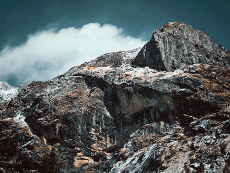 Rocky mountain peak with patches of snow under a partly cloudy sky, showing rugged textures and dramatic contrast between the dark rock and lighter highlights.