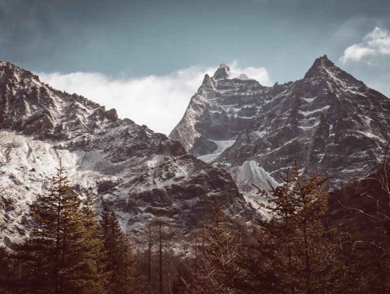 Snow-dusted, rugged mountain peaks rise above a forest of evergreen trees under a partly cloudy sky, creating a dramatic and scenic landscape.