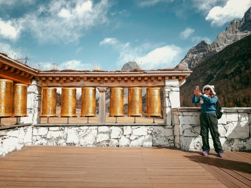 A person in a blue jacket stands on a wooden platform next to large golden prayer wheels, with snowy mountains and a partly cloudy sky in the background.