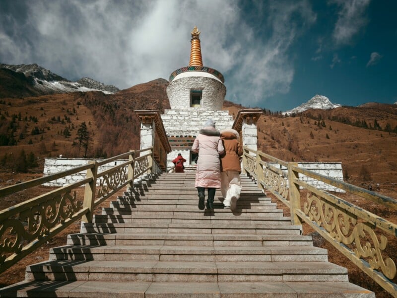 Two people in winter coats ascend a stone staircase towards a stupa with a colorful spire, set against a mountainous, partly cloudy landscape. The scene is peaceful and atmospheric.