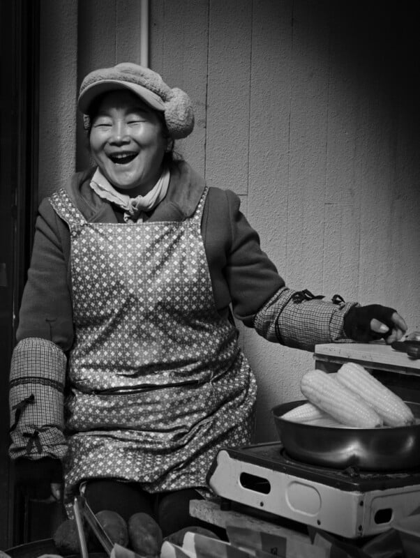 A woman wearing a patterned apron, hat, and earmuffs smiles joyfully while sitting next to a pan of corn and crates. The image is in black and white.