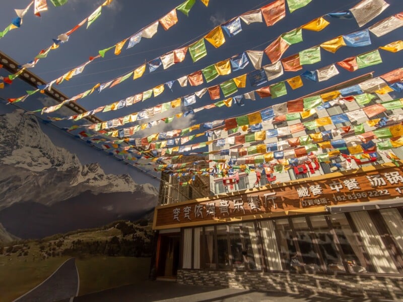 Colorful prayer flags are strung across an outdoor area with a mountainscape mural on one wall and a building with Chinese signage and wooden accents in the background, under a sunny sky.