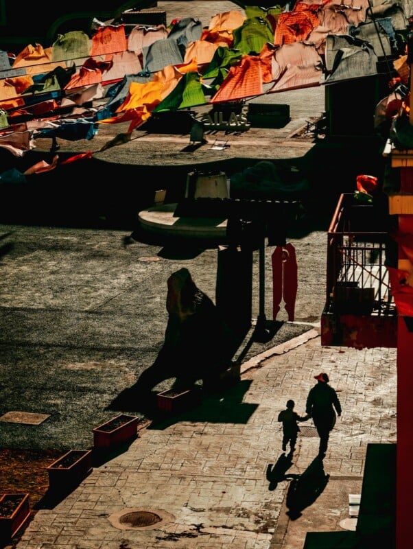 Two people, an adult and a child, walk hand in hand, casting long shadows on a sunlit street. Above them, rows of colorful fabric or flags hang, creating a vibrant, lively atmosphere.