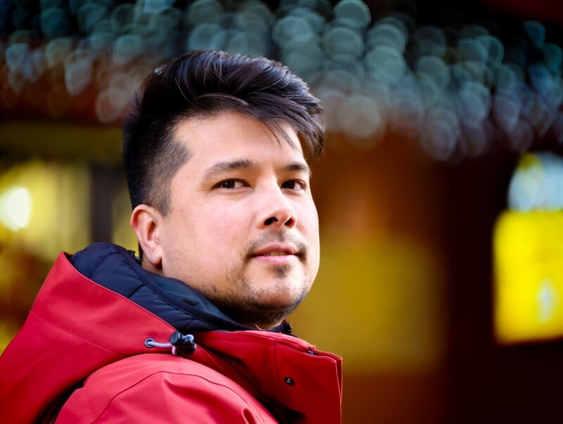 A man with short dark hair and light facial hair wearing a red jacket looks at the camera. The background is blurred with circular light bokeh and warm colors.