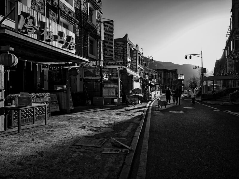 A black-and-white photo of a quiet street with traditional-style buildings, a few people and a dog walking along the sidewalk, streetlights, and mountains visible in the background under bright sunlight.