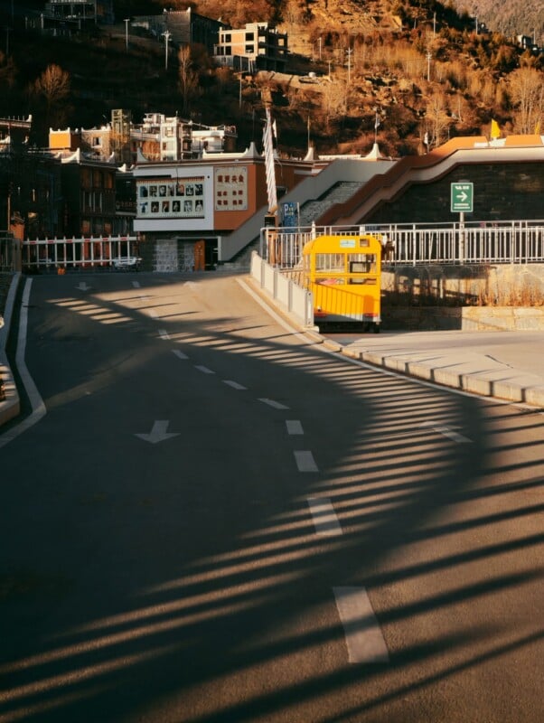 A yellow school bus drives along a curved road toward a gated entrance in a mountainous area, with shadows from the fence creating stripes on the asphalt. Buildings and dry trees are visible in the background.