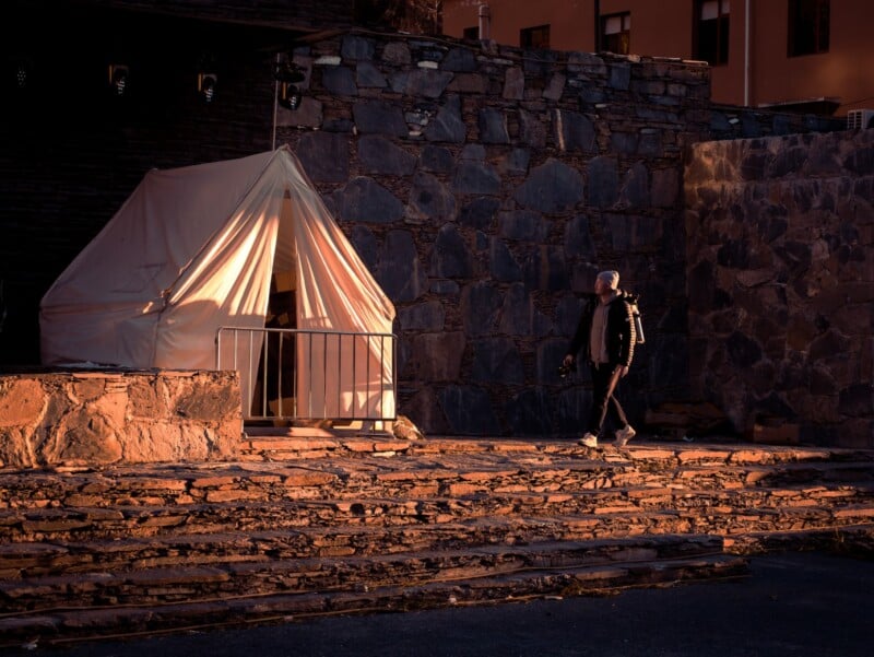 A person walks past a white tent set against a stone wall at sunset, casting long shadows and warm light across the stone steps and ground.