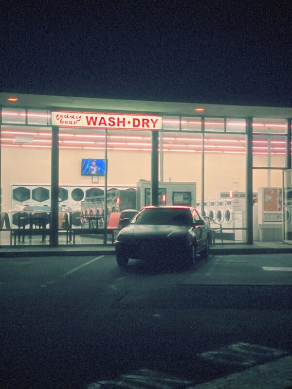 Por la noche, un coche oscuro estacionó frente a una lavandería muy iluminada. El cartel de arriba dice "Oso de peluche limpieza·secado," A través de las ventanas de cristal se pueden ver hileras de lavadoras y sillas.