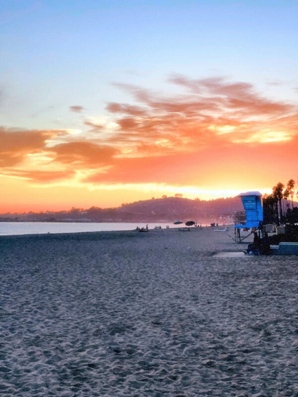 La playa al atardecer, el cielo colorido, el océano en calma, las colinas a lo lejos y una torre de salvavidas azul a la derecha. Al fondo se pueden ver siluetas de personas y palmeras.