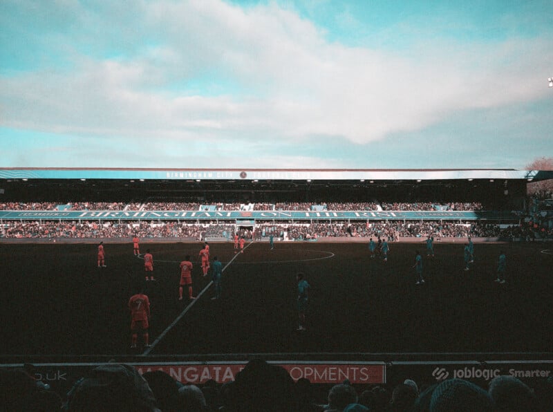 Two soccer teams stand on the field at Birmingham City’s St. Andrew's stadium, preparing for kickoff, as spectators watch from the stands under a partly cloudy sky.