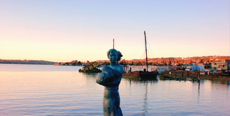 Una estatua de una mujer sosteniendo un objeto se encuentra junto a la orilla del agua al atardecer, con barcos, un muelle y una costa bordeada de edificios y árboles al fondo.