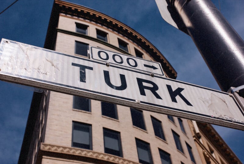El cartel de la calle dice "turco" y "000 →" El fondo en negrita es un edificio beige con varias ventanas y un cielo azul.