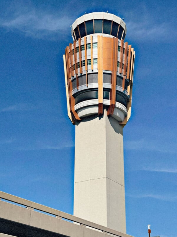 Una moderna torre de control de aeropuerto con una base cilíndrica y una estructura superior única con paneles naranjas y grises contra el cielo azul claro.