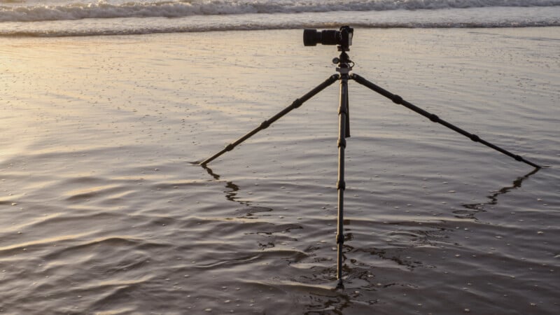 A camera mounted on a tripod stands in shallow water at the beach, with gentle waves and the ocean visible in the background during sunset.