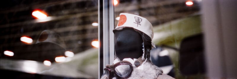 A mannequin dressed in vintage pilot gear, including a white cap with Japanese writing and a red circle, is displayed behind glass with blurred lights and an aircraft in the background.