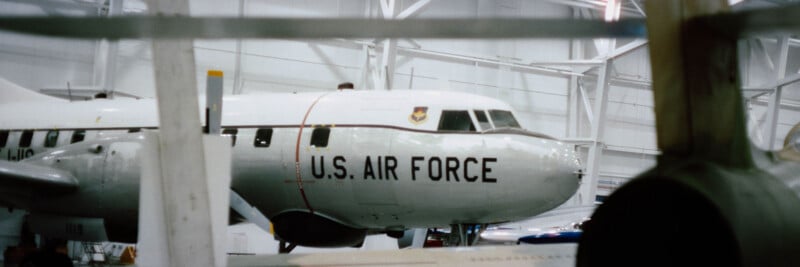 A U.S. Air Force aircraft is displayed inside a hangar, partially obscured by structural beams and another plane, with white walls and bright lighting in the background.