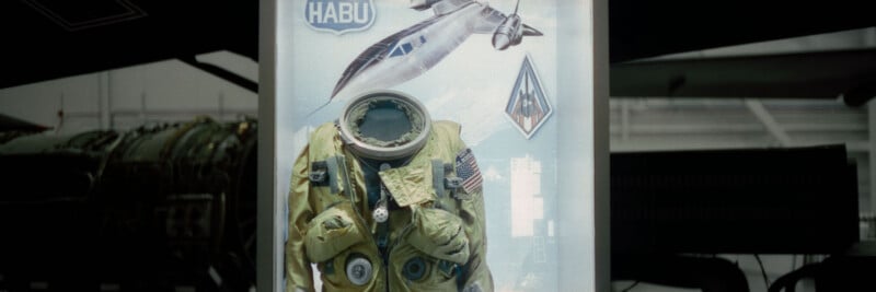 A display case featuring a vintage flight suit with patches, including a US flag, in front of a poster showing a jet aircraft and the word "HABU." The background is dim and industrial.