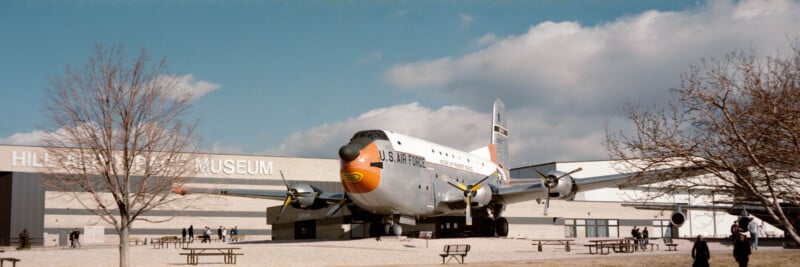 A large vintage U.S. Air Force airplane is displayed outside the Hill Aerospace Museum. People walk nearby, and leafless trees border the scene under a partly cloudy sky.