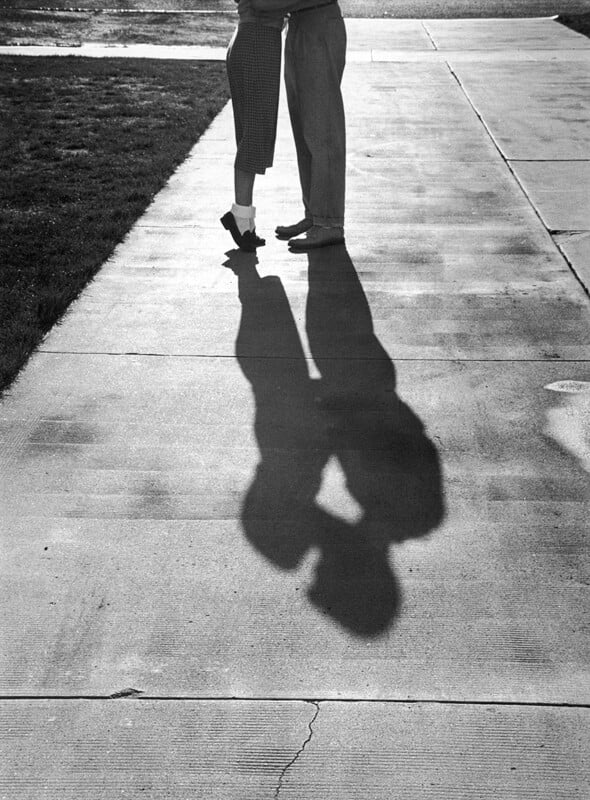 A couple stands close together on a sidewalk, their upper bodies out of frame. The sunlight casts a clear shadow of them embracing or possibly kissing on the pavement. Grass borders the sidewalk on one side.