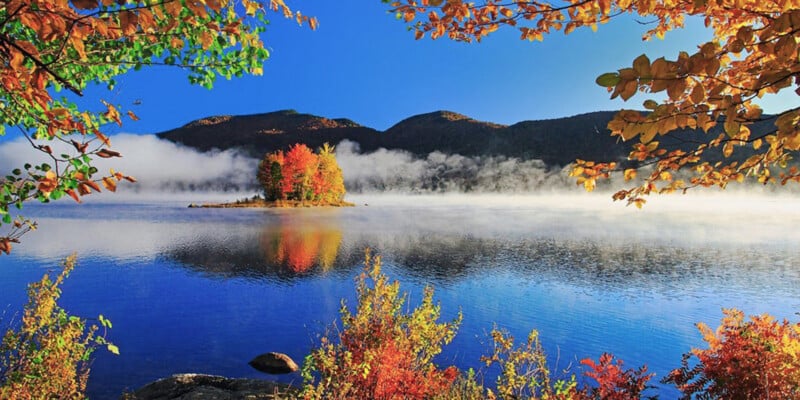 A small island with colorful autumn trees is reflected in a calm lake, surrounded by mist and mountains, with vibrant fall foliage framing the scene under a clear blue sky.