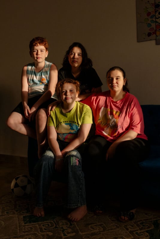 Four children sit closely together in a warmly lit room, some smiling slightly. The group is casually dressed, with a soccer ball on the floor and a patterned rug underneath them. The background wall is plain with partial artwork visible.