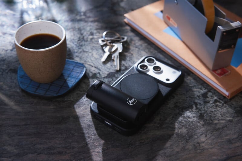 A cup of black coffee sits on a coaster beside a smartphone with a camera attachment, a set of keys, a notebook, and a tape dispenser on a marble surface.