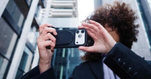 A person with curly hair holds up a smartphone horizontally, taking a photo or video, while standing outdoors among modern tall buildings.