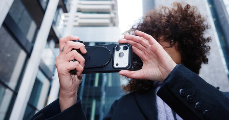 A person with curly hair holds up a smartphone horizontally, taking a photo or video, while standing outdoors among modern tall buildings.
