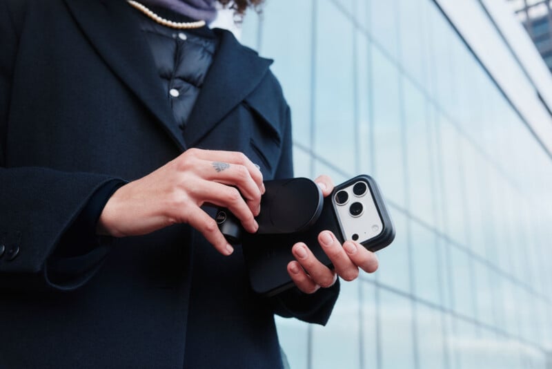 A person wearing a black coat holds a smartphone and attaches a circular device to its back, standing in front of a glass building.