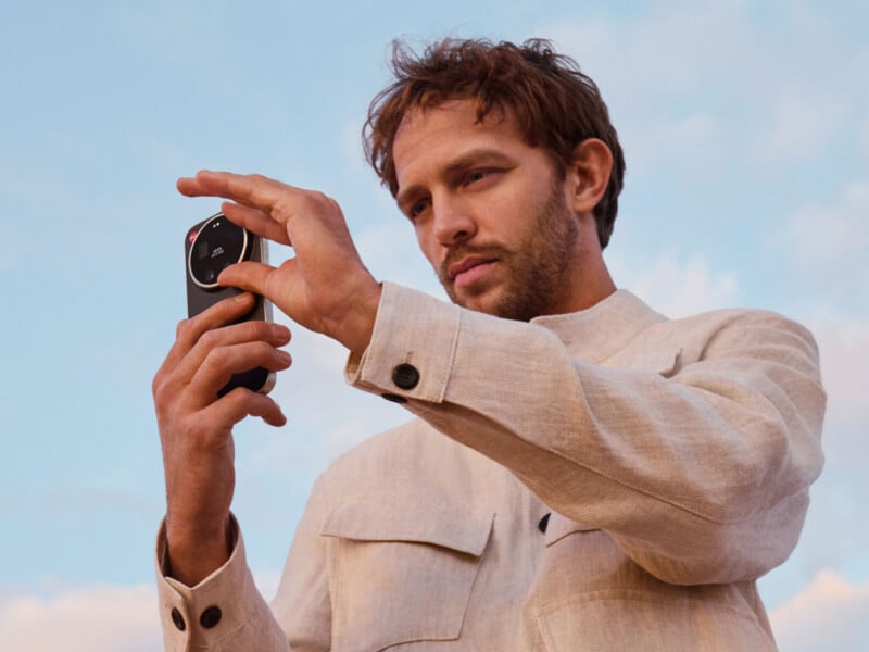 A man wearing a beige jacket stands outdoors, holding and pointing at a small black electronic device with buttons, against a blue sky with clouds.