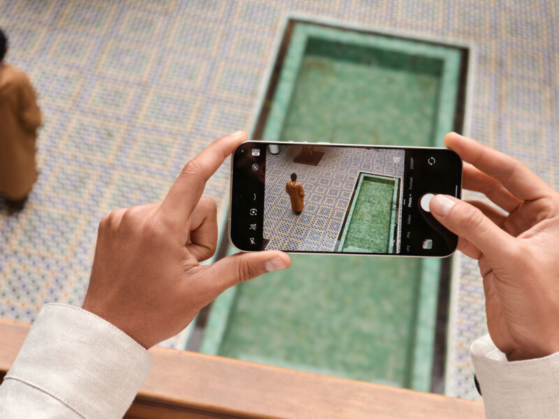 A person holds a smartphone, taking a photo of another person standing near a rectangular tiled pool with a patterned floor. The pool and person are viewed from above.
