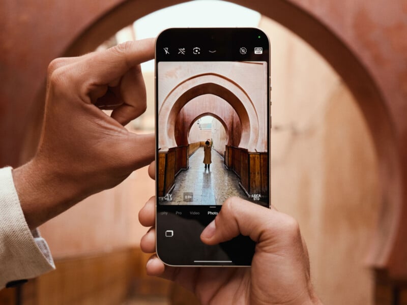 A person takes a photo with a smartphone, capturing another person standing under a series of arched walkways with warm, brown-toned walls in the background.