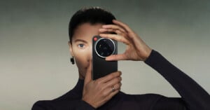 A woman with slicked-back hair holds a smartphone with a large camera lens in front of one eye, as if taking a photo. She wears a dark outfit, statement earring, and a ring, against a neutral background.