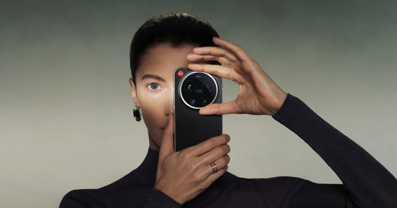 A woman with slicked-back hair holds a smartphone with a large camera lens in front of one eye, as if taking a photo. She wears a dark outfit, statement earring, and a ring, against a neutral background.