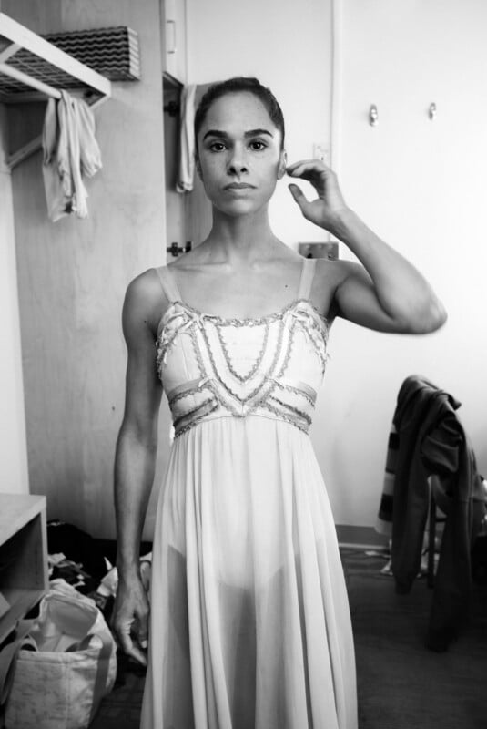 Una mujer con un traje de ballet sin mangas se encuentra en un camerino y mira directamente a la cámara. Se alisó el cabello con una mano. Al fondo se ven claramente ropa y bolsos. Las fotos están en blanco y negro.