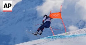 A skier wearing a racing suit and helmet speeds downhill past an orange Olympic gate flag on a snowy slope, with mountains and clouds in the background. The AP logo appears in the top left corner.