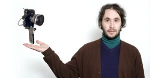 A man with curly hair and a beard, wearing a turtleneck and a brown sweater, holds out his hand as an old-fashioned camera appears to float above his palm against a plain white background.