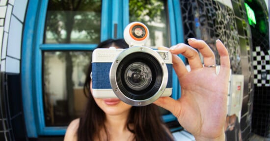A person holds a large-lens camera up to their face, obscuring their features. The background shows a blue window, green trees reflected in the glass, and a black-and-white tiled wall.