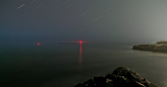 Long-exposure night photo of a calm sea with star trails in the sky, two red navigation lights reflecting on the water, and dark rocky shorelines visible in the foreground and to the right.