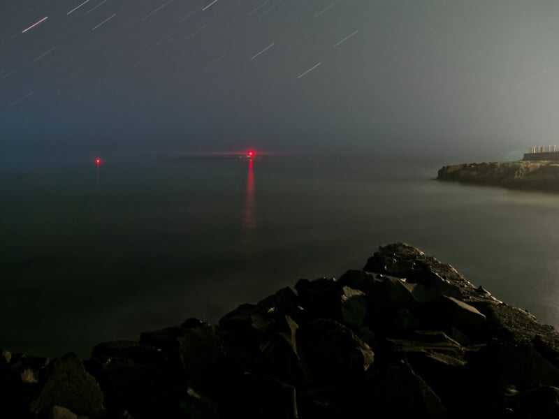 Costa rocosa por la noche, dos luces de navegación rojas reflejadas en las aguas tranquilas; Se pueden ver rastros de estrellas en el cielo.