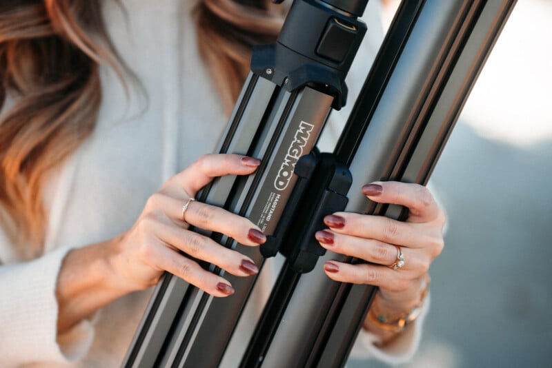 Un hombre con cabello largo y uñas pintadas sostiene un trípode Magco doblado, mostrando la etiqueta y el cierre de la marca, mientras viste un suéter color crema y anillos dorados.