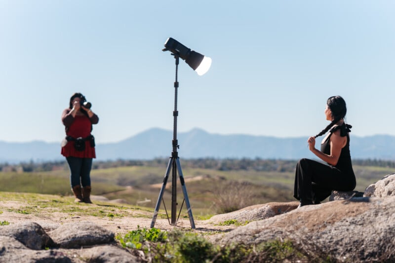 Un fotógrafo utiliza una gran luz fotográfica montada en un trípode para fotografiar a una mujer sentada al aire libre. Están situadas sobre un paisaje rocoso con montañas y el cielo azul de fondo.