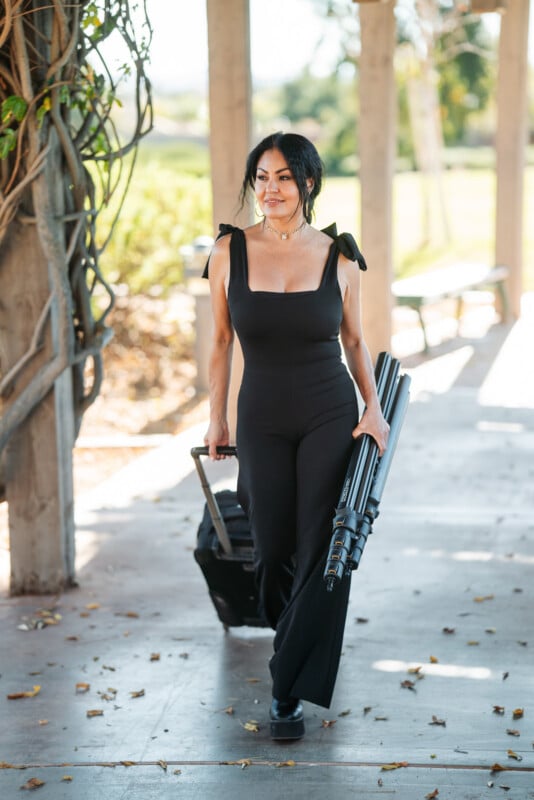 Una mujer vestida con un mono negro salió, sosteniendo una maleta en una mano y un trípode doblado en la otra. Ella sonríe, rodeada de vegetación y pilares de madera.