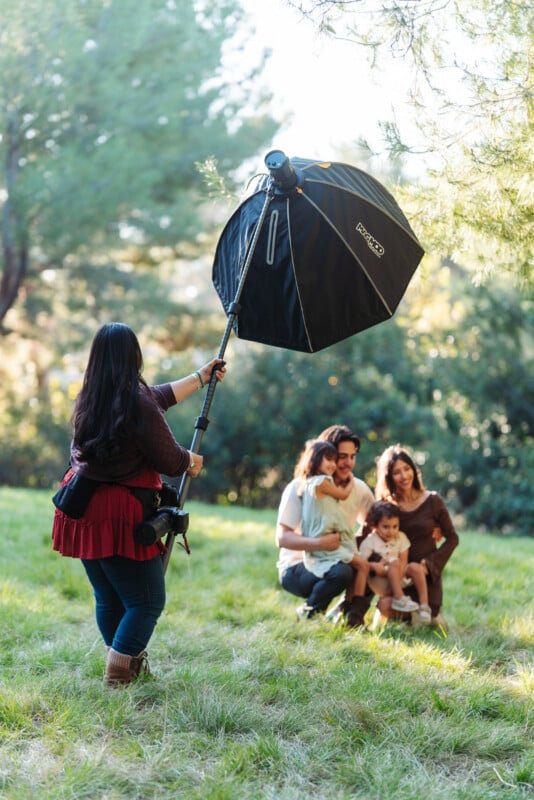 Una mujer sostiene un gran difusor fotográfico y una familia de cuatro personas posa sobre el césped en un soleado parque al aire libre. Árboles y vegetación al fondo.